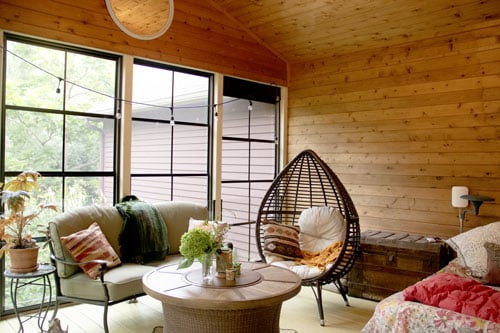 Cedar-paneled porch with a daybed, egg chair, and sofa beside tall black-framed porch windows