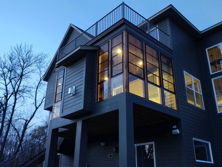 Porch Windows from exterior of dark grey home lit from the inside with deck above the porch